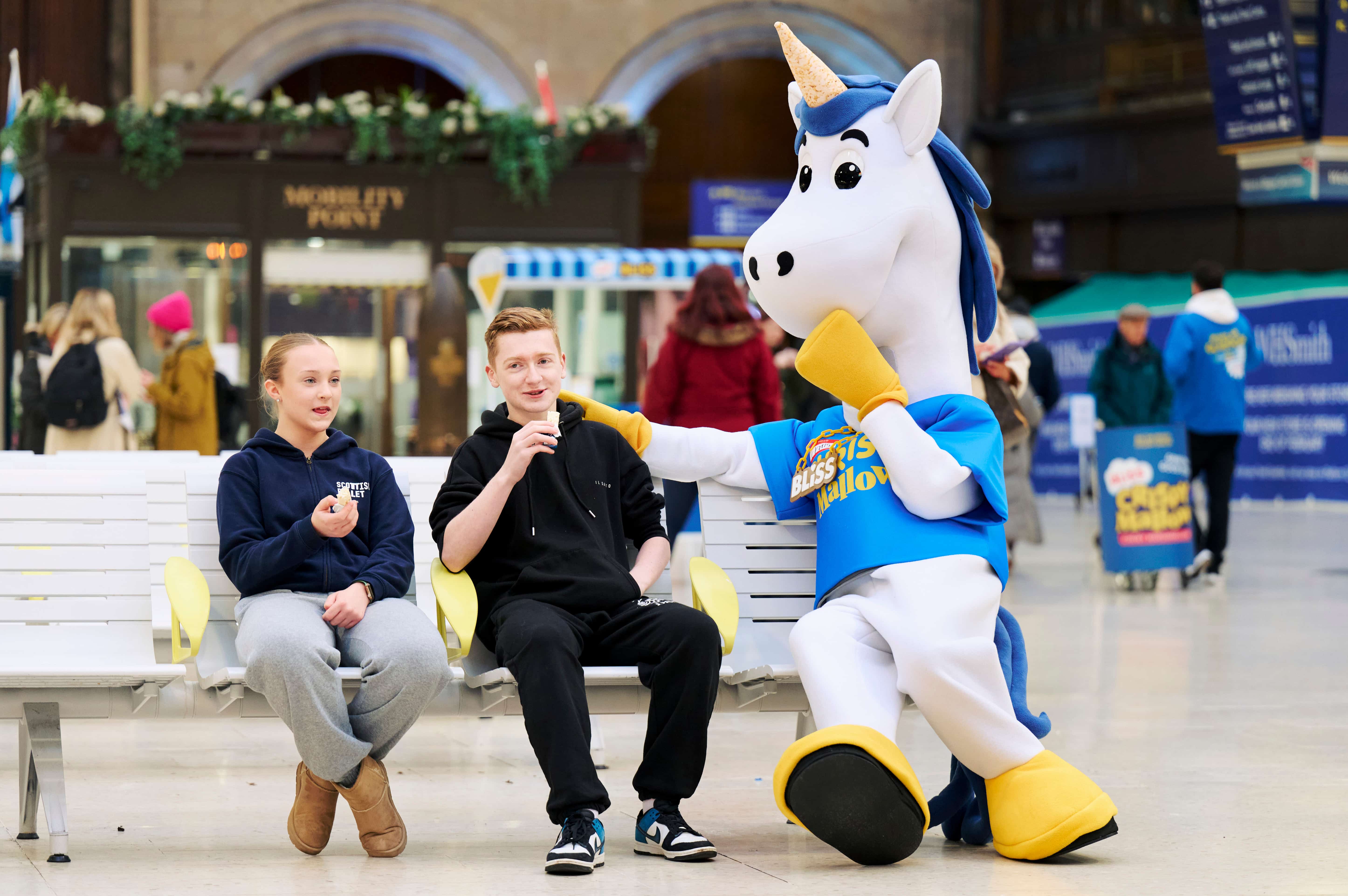 Two kids on a bench sitting next to a unicorn mascot in a train station