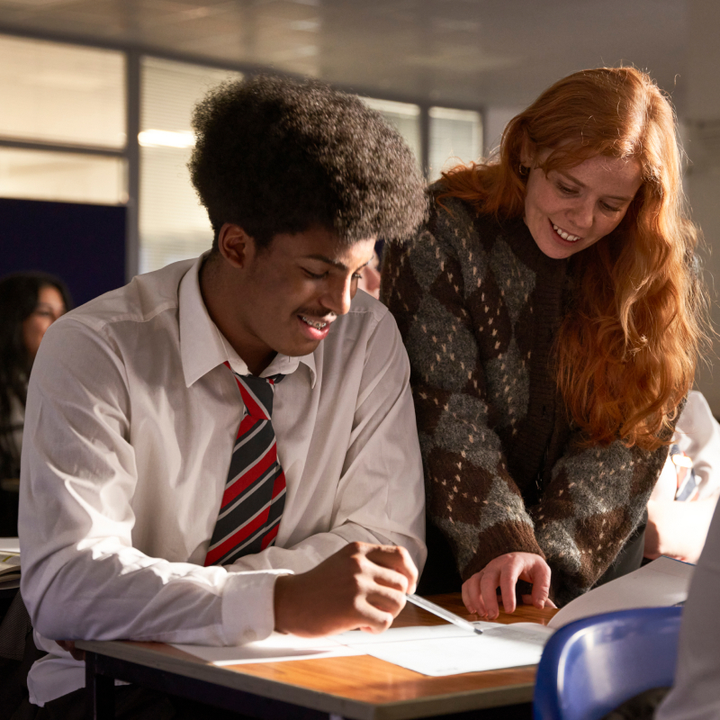 Pupil sat in class with female teacher standing over him, helping him with lesson